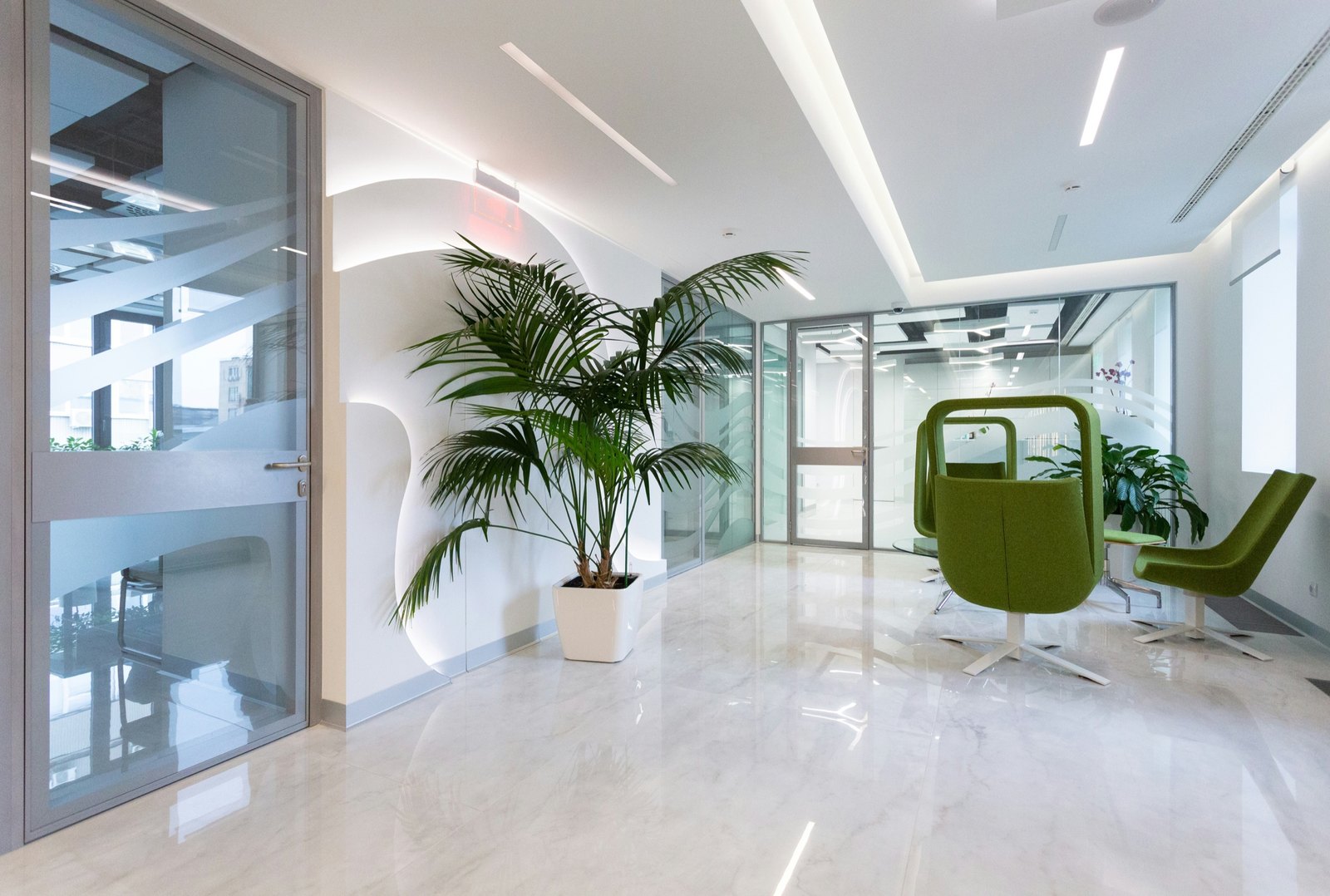 Bright clinic reception with a marble floor, a potted palm, green armchairs and glass partitions.